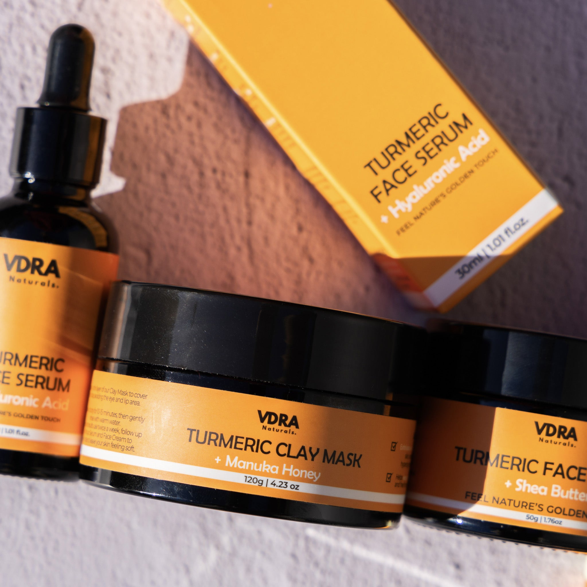 VDRA Naturals skincare products including a tumreric serum, clay mask, and face serum on a textured surface.