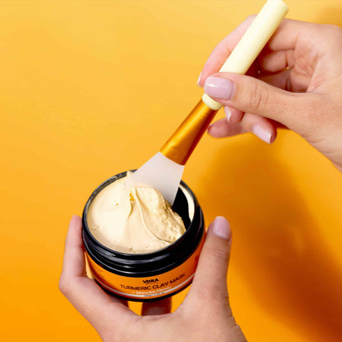 Person scooping the Turmeric Clay Mask with the applicator tool, against an orange background.