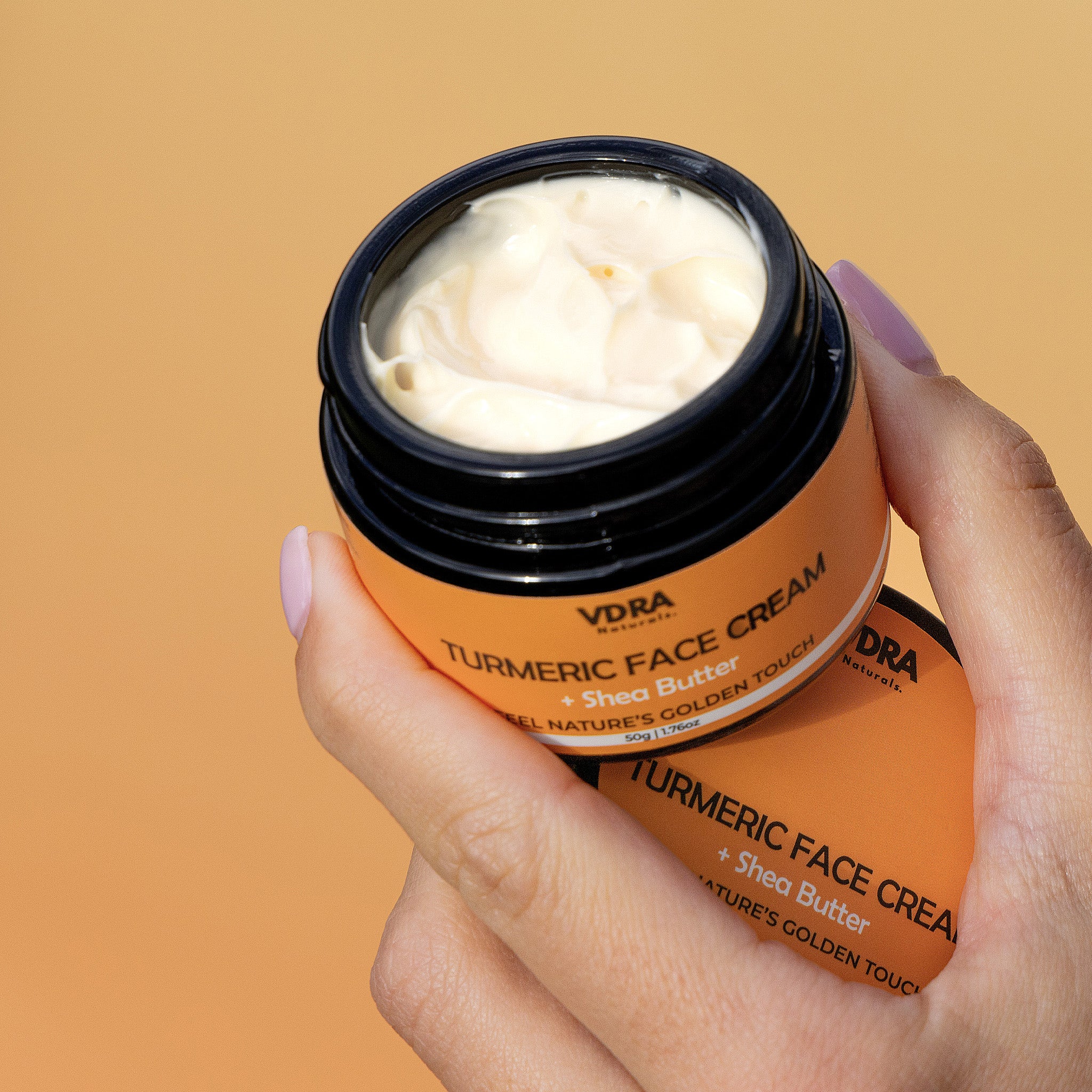Hand holding an open jar of VDRA Turmeric Face Cream with a beige background