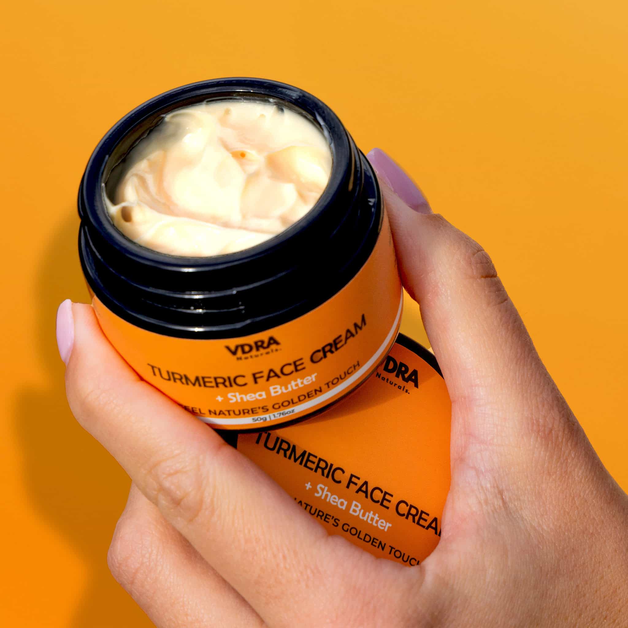 Orange jar of VDRA Turmeric Face Cream held by a hand against an orange background, highlighting the soft, creamy texture of the cream. 
