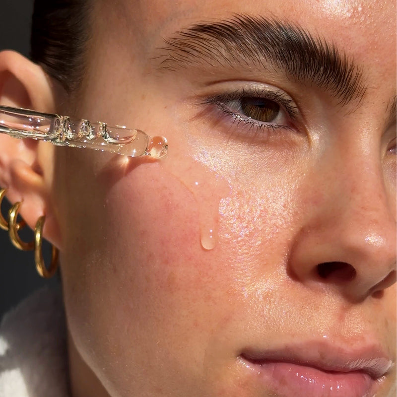 Close-up of a person's face with a droplet of the turmeric face serum on their cheek, wearing gold hoop earrings.